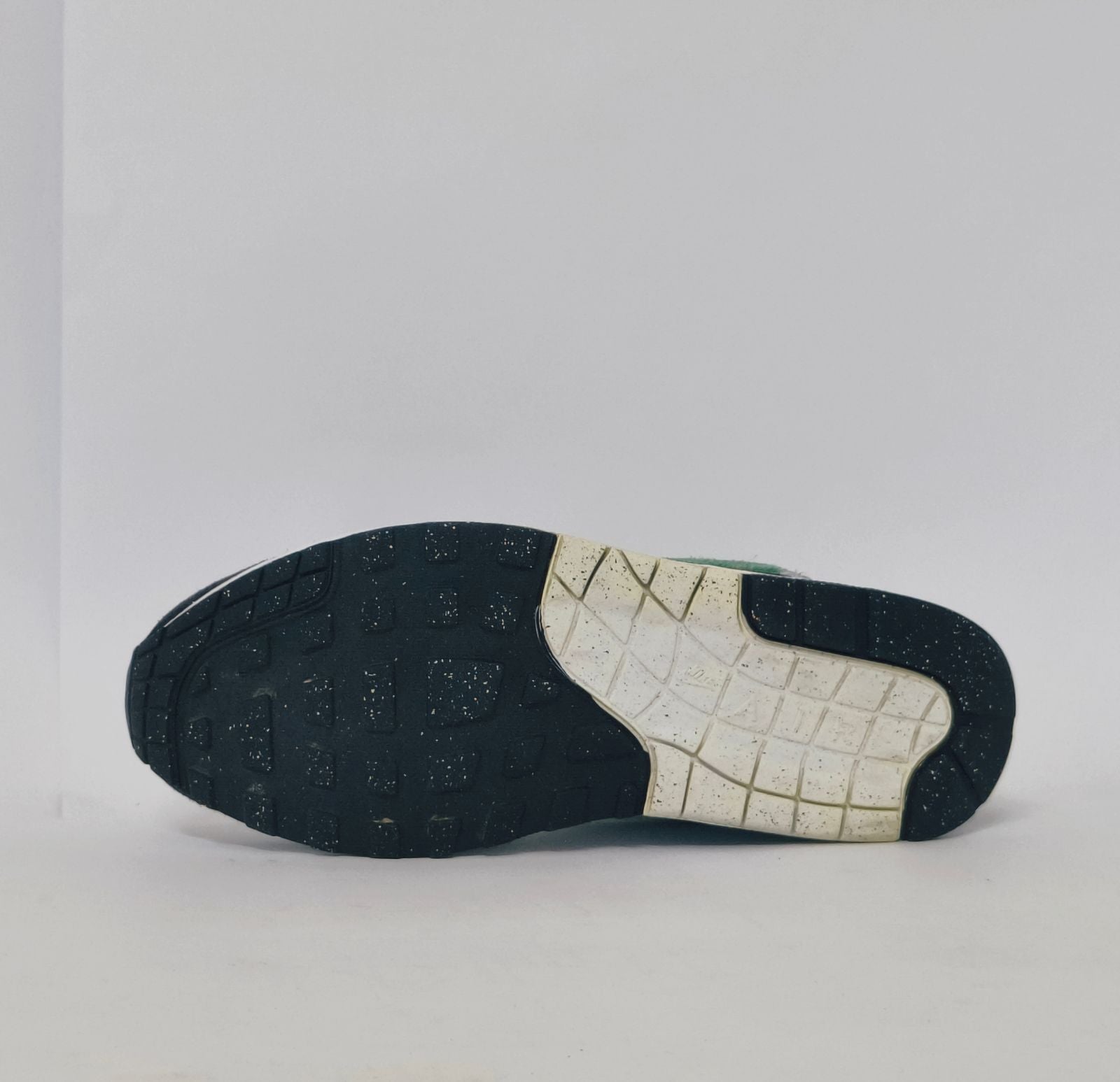 Nike Air Max 1 sneaker featuring a grey upper, black mudguard, and a green suede Swoosh with a visible air bubble in the white midsole.