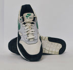 Nike Air Max 1 sneaker featuring a grey upper, black mudguard, and a green suede Swoosh with a visible air bubble in the white midsole.