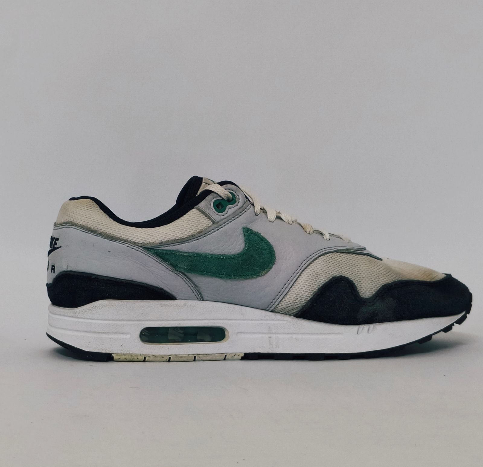 Nike Air Max 1 sneaker featuring a grey upper, black mudguard, and a green suede Swoosh with a visible air bubble in the white midsole.