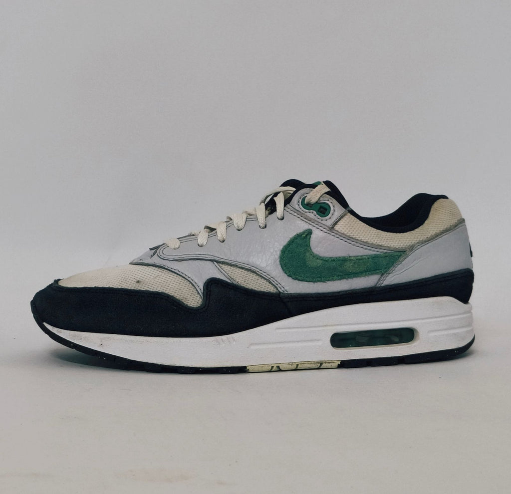 Nike Air Max 1 sneaker featuring a grey upper, black mudguard, and a green suede Swoosh with a visible air bubble in the white midsole.