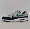 Nike Air Max 1 sneaker featuring a grey upper, black mudguard, and a green suede Swoosh with a visible air bubble in the white midsole.