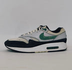 Nike Air Max 1 sneaker featuring a grey upper, black mudguard, and a green suede Swoosh with a visible air bubble in the white midsole.
