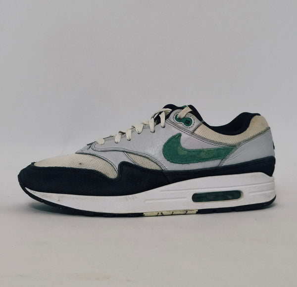 Nike Air Max 1 sneaker featuring a grey upper, black mudguard, and a green suede Swoosh with a visible air bubble in the white midsole.