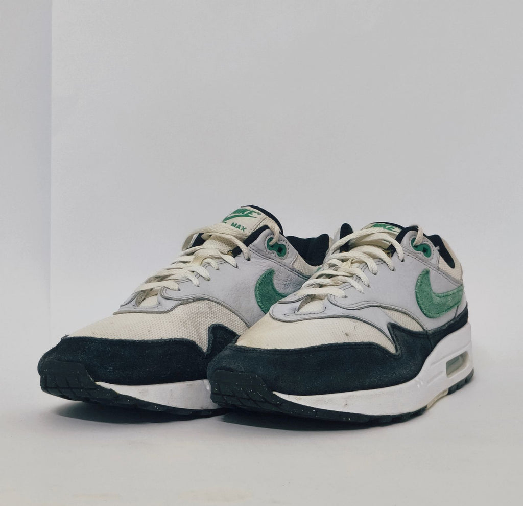 Nike Air Max 1 sneaker featuring a grey upper, black mudguard, and a green suede Swoosh with a visible air bubble in the white midsole.
