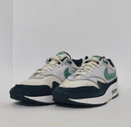 Nike Air Max 1 sneaker featuring a grey upper, black mudguard, and a green suede Swoosh with a visible air bubble in the white midsole.