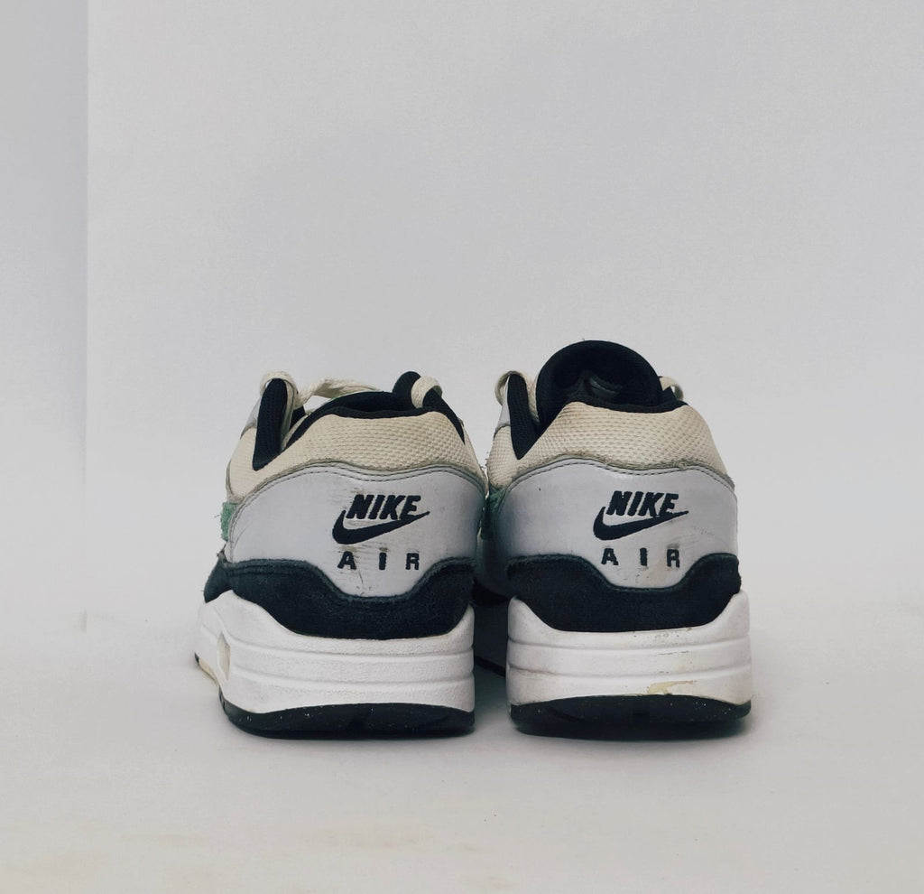 Nike Air Max 1 sneaker featuring a grey upper, black mudguard, and a green suede Swoosh with a visible air bubble in the white midsole.