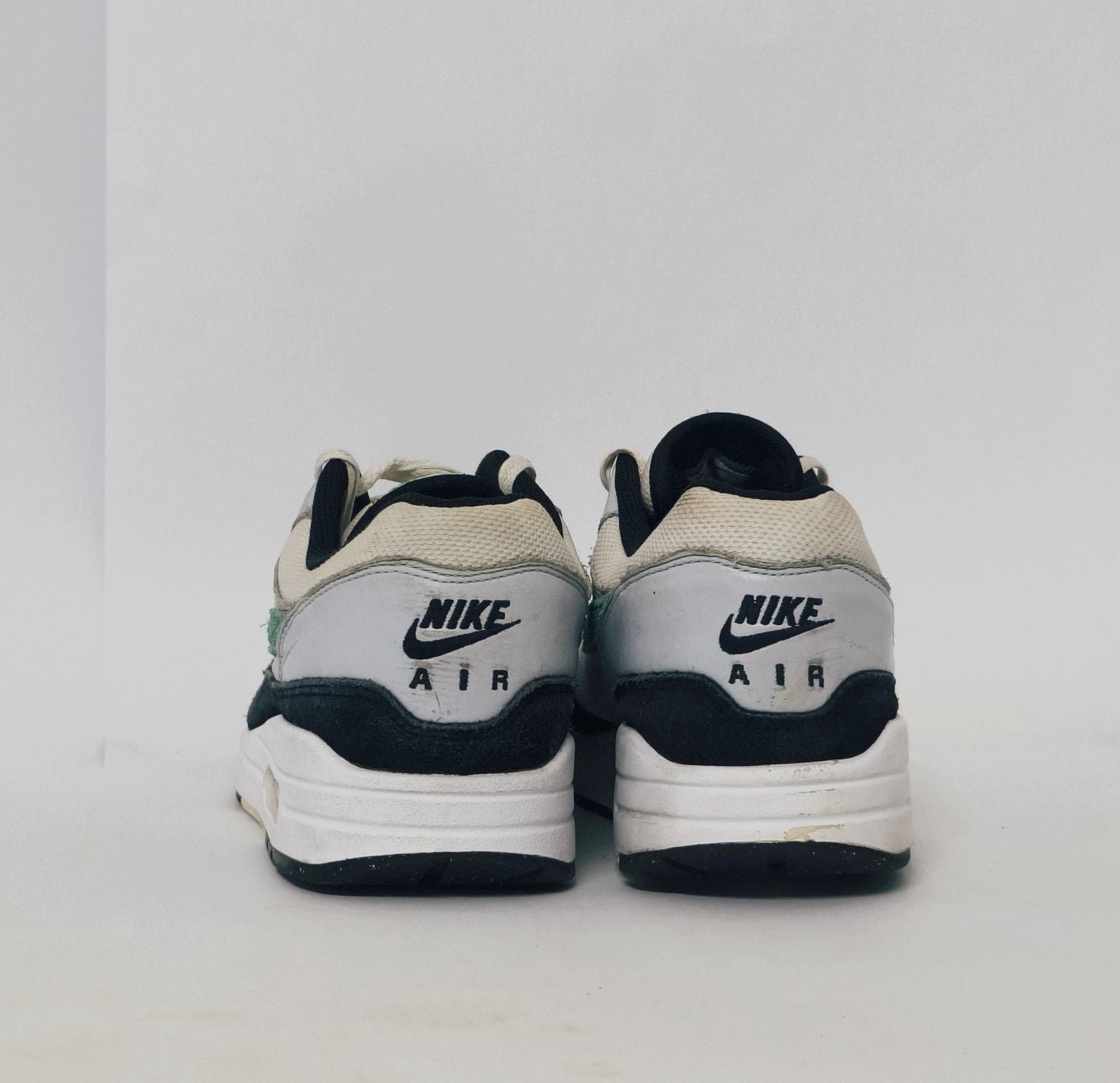 Nike Air Max 1 sneaker featuring a grey upper, black mudguard, and a green suede Swoosh with a visible air bubble in the white midsole.