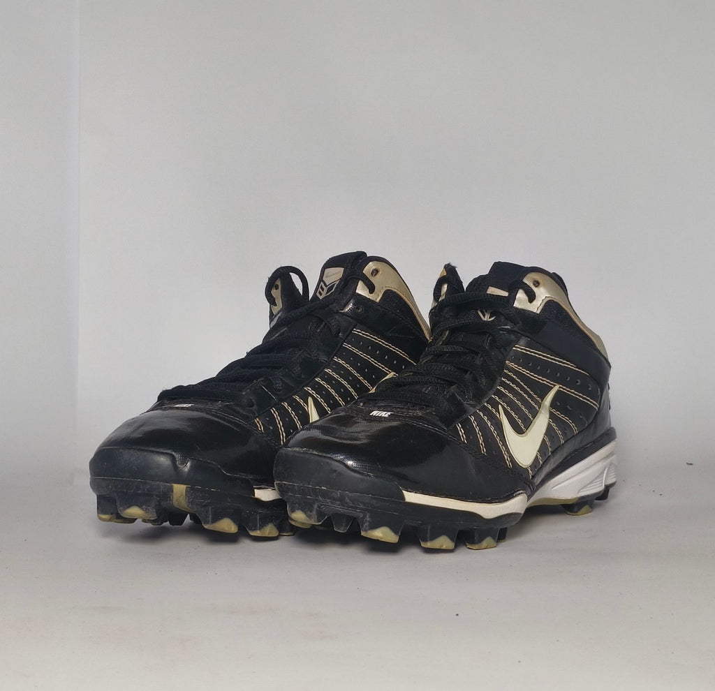 black Nike Huarache baseball cleat featuring a white Swoosh, gold heel accents, and a white midsole with a cleated outsole.