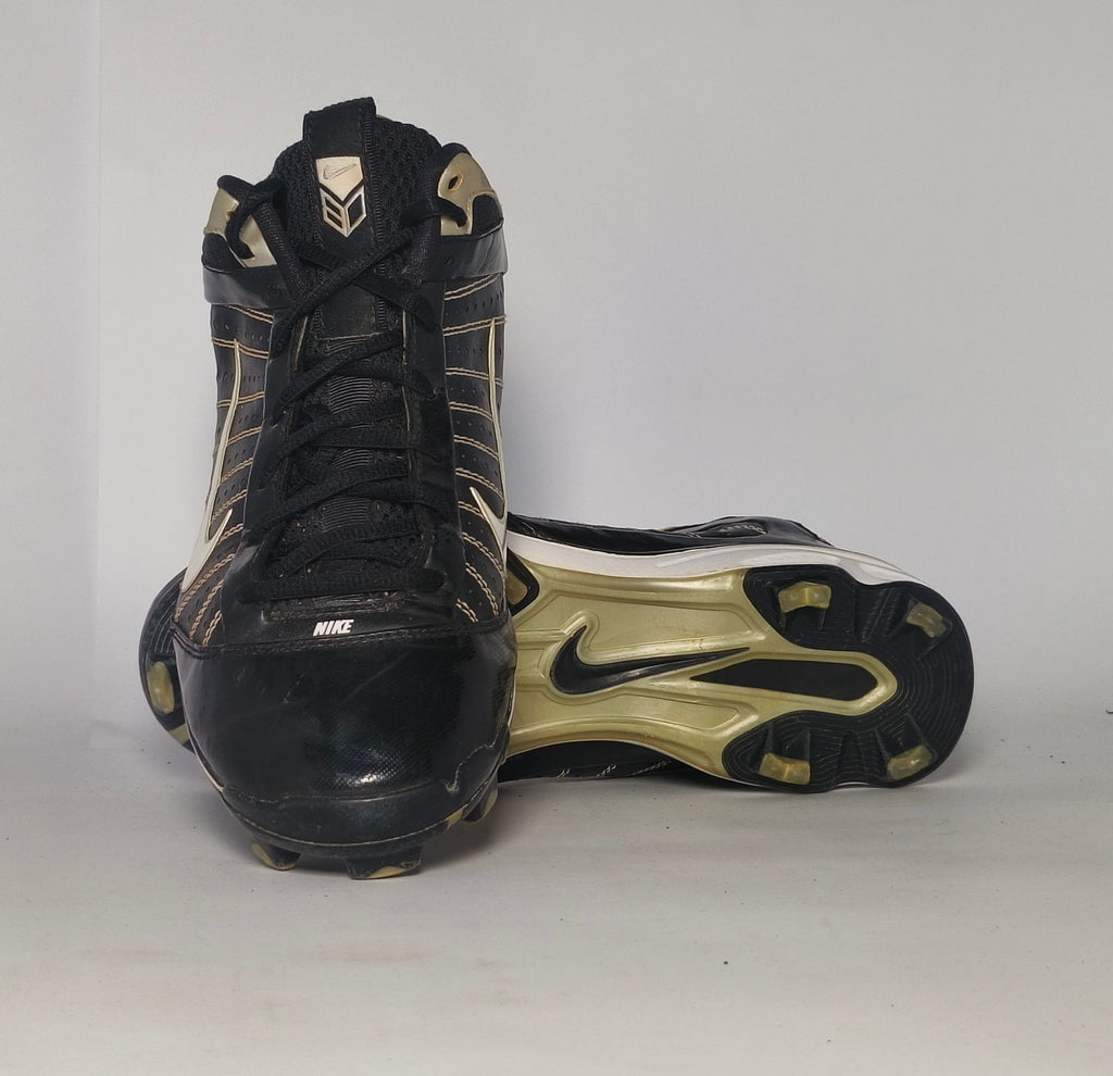 black Nike Huarache baseball cleat featuring a white Swoosh, gold heel accents, and a white midsole with a cleated outsole.