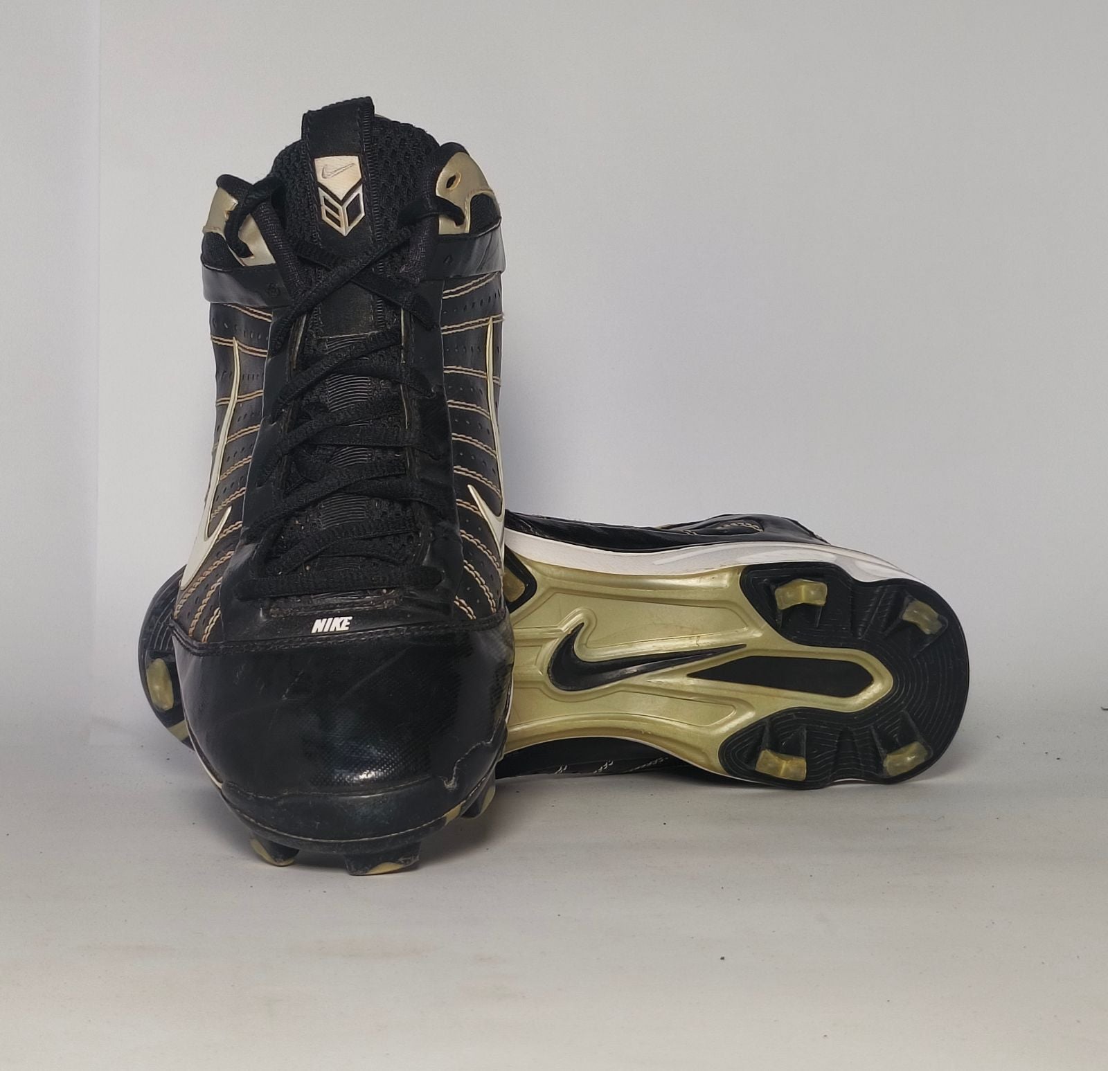 black Nike Huarache baseball cleat featuring a white Swoosh, gold heel accents, and a white midsole with a cleated outsole.