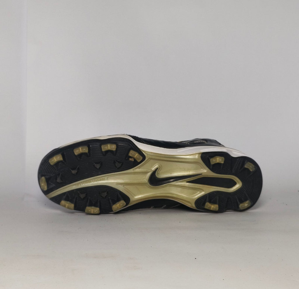 black Nike Huarache baseball cleat featuring a white Swoosh, gold heel accents, and a white midsole with a cleated outsole.