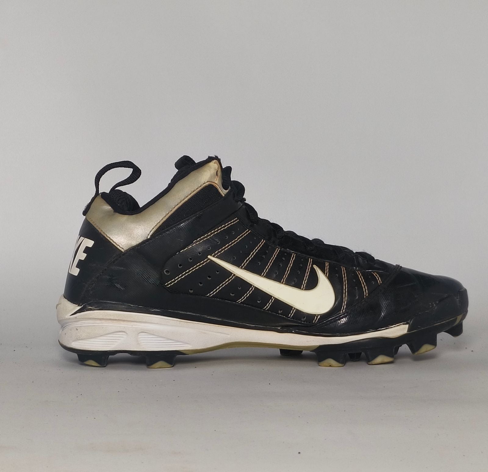 black Nike Huarache baseball cleat featuring a white Swoosh, gold heel accents, and a white midsole with a cleated outsole.